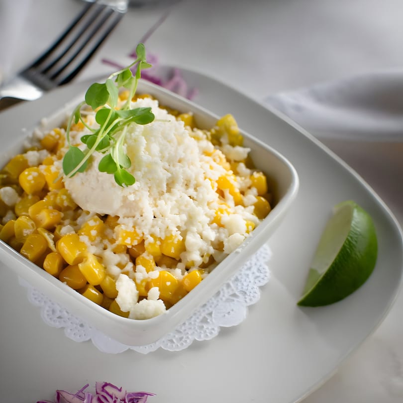 Mexican street corn (Esquites) in a white bowl, topped with cheese, crema, and microgreens, with a lime wedge.