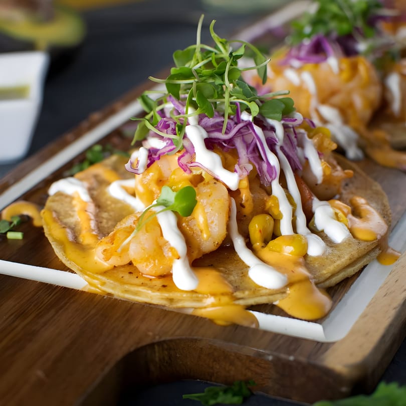 Close-up of shrimp tacos with red cabbage, microgreens, and creamy sauces on a wooden tray.
