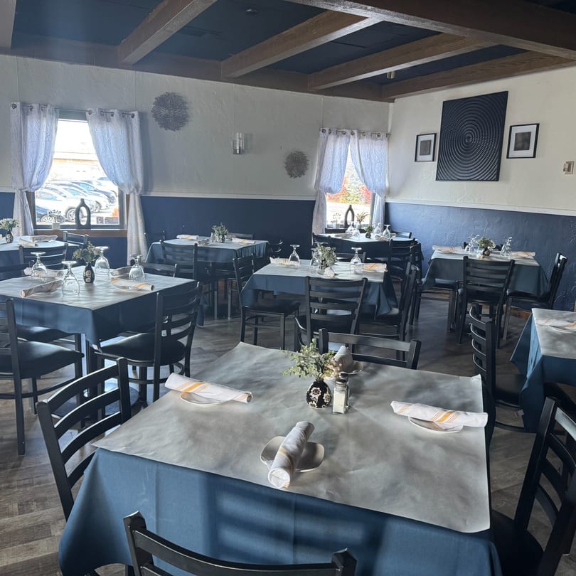 Interior of Tavos at the Grove, showing dining tables with blue tablecloths, black chairs, and window views.