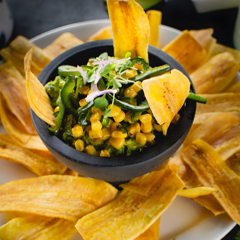 Guacamole with corn and poblano peppers, garnished with microgreens, surrounded by plantain chips.