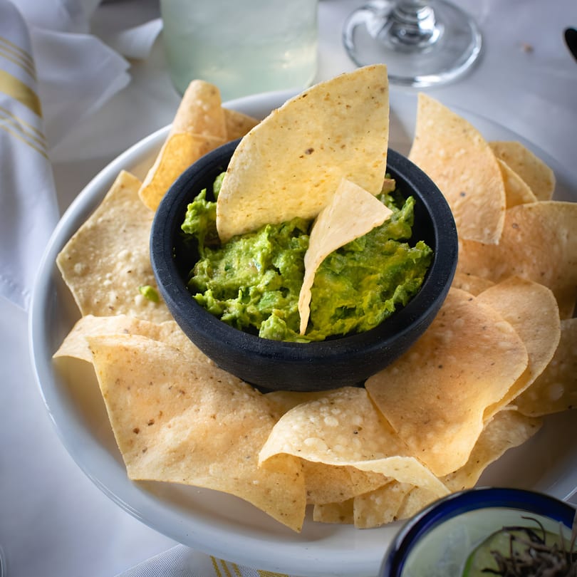 A black bowl of vibrant green guacamole surrounded by crispy tortilla chips on a white plate.