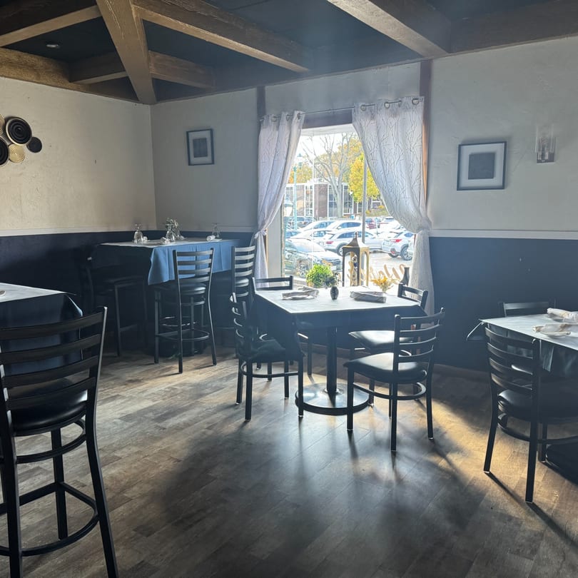 Interior of Tavos at the Grove, showing dining tables with blue tablecloths and a window view.